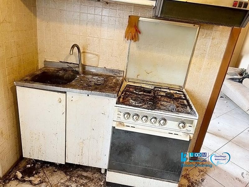 Una cucina sporca e trascurata con un lavandino macchiato, armadietti sudici e un piano cottura unto e arrugginito. Un guanto di gomma arancione è drappeggiato sul retro della lavagna: una vera bozza automatica di sporcizia, dove anche il pavimento e le pareti sono macchiati e sporchi.