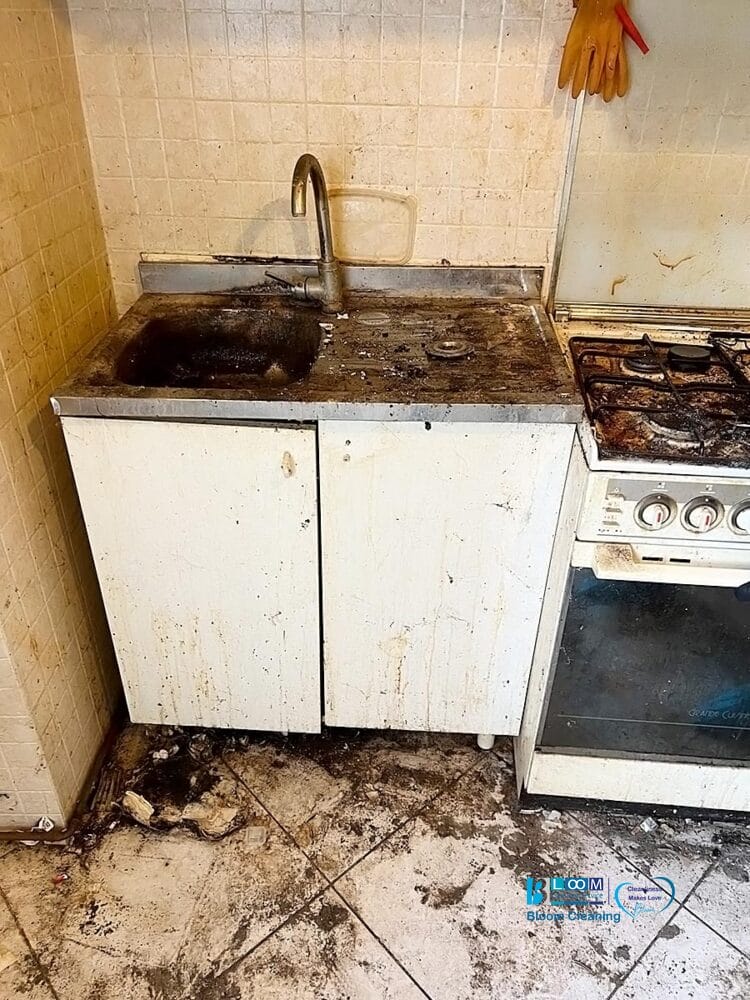 Una cucina sporca con lavello macchiato, banconi sporchi e armadietti sudici. Il pavimento è coperto di sporcizia e anche i fornelli sono molto sporchi. Sopra il lavello è appeso un guanto di gomma, vera bozza automatica di incuria.