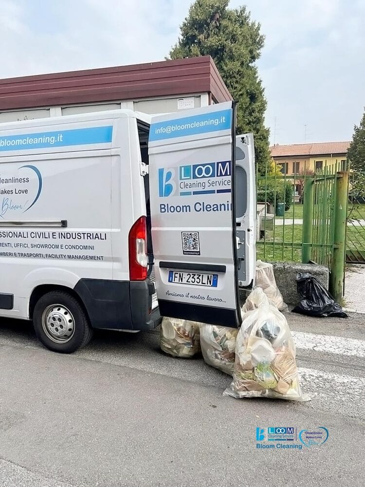 Un furgone bianco per le pulizie è parcheggiato con la porta posteriore aperta. Diversi grandi sacchi pieni di rifiuti sono posizionati accanto al furgone sulla strada, vicino a una recinzione verde e ad alcuni alberi, come parte di un'operazione di pulizia automatica di Bozza.