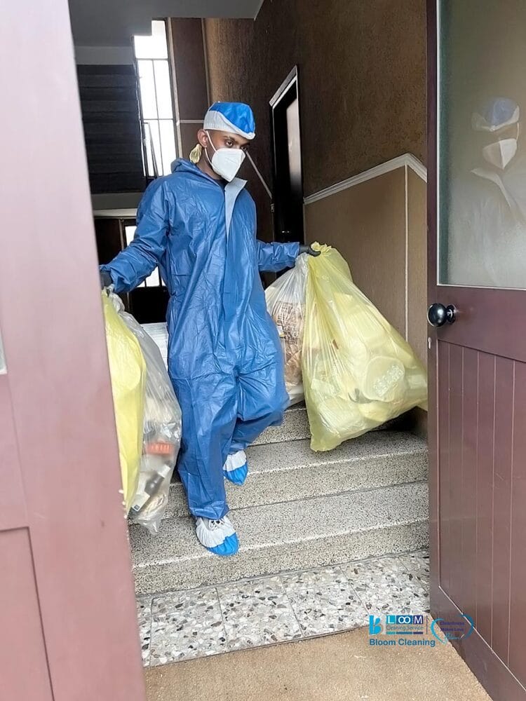 Una persona con tuta protettiva blu, berretto, maschera e copriscarpe trasporta due grandi sacchi gialli per la spazzatura lungo una scala all'interno di un edificio. La porta è aperta e "Bozza automatica" è visibile su un'insegna vicina.