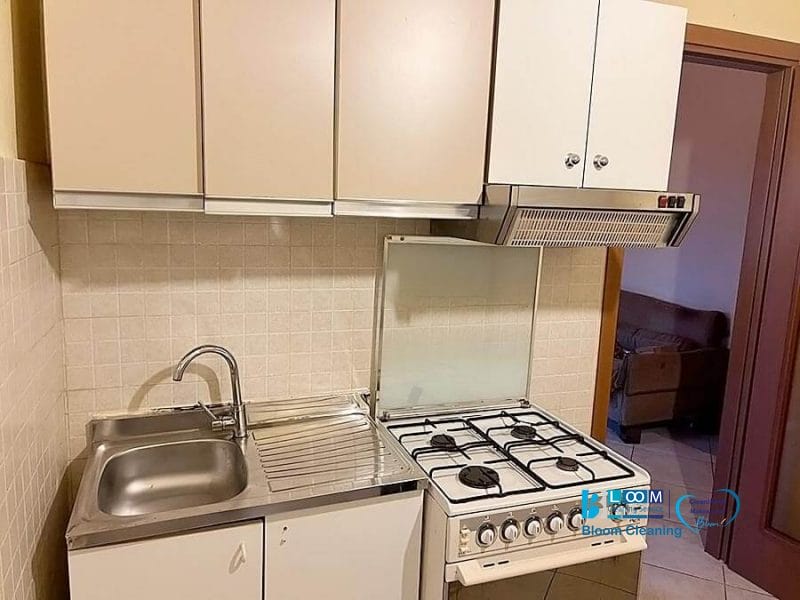 Una piccola cucina con lavello in acciaio inox, sgocciolatoio, pensili e un fornello a gas a quattro fuochi. Un forno e una cappa completano lo spazio. Una porta aperta rivela parte di una stanza con divano marrone e arredamento automatico Bozza.