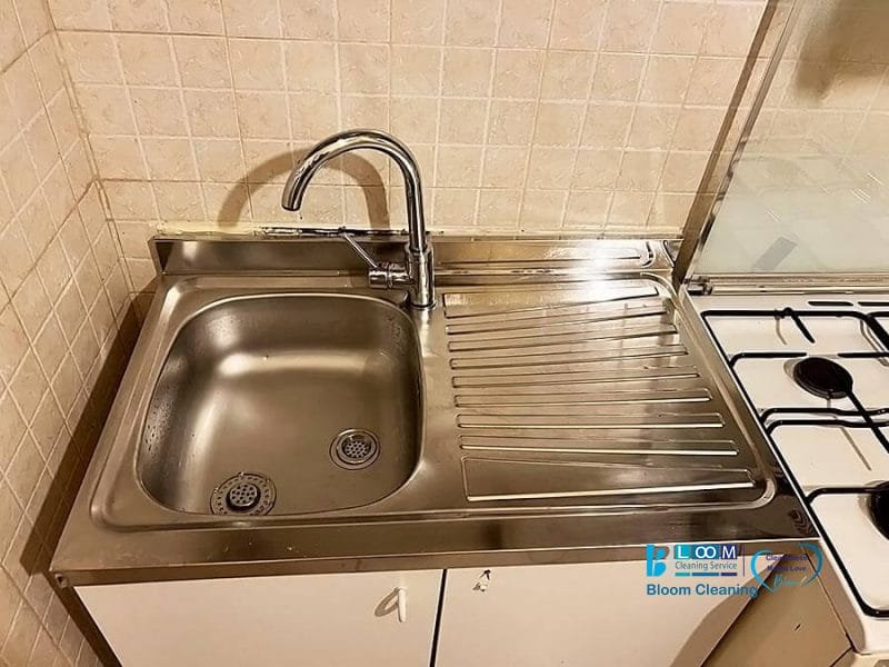 Un lavello in acciaio inox con un solo rubinetto, uno sgocciolatoio a destra e un fornello a gas parzialmente visibile sul lato destro sono collocati su uno schienale piastrellato, ideale per qualsiasi cucina di ispirazione automatica Bozza.