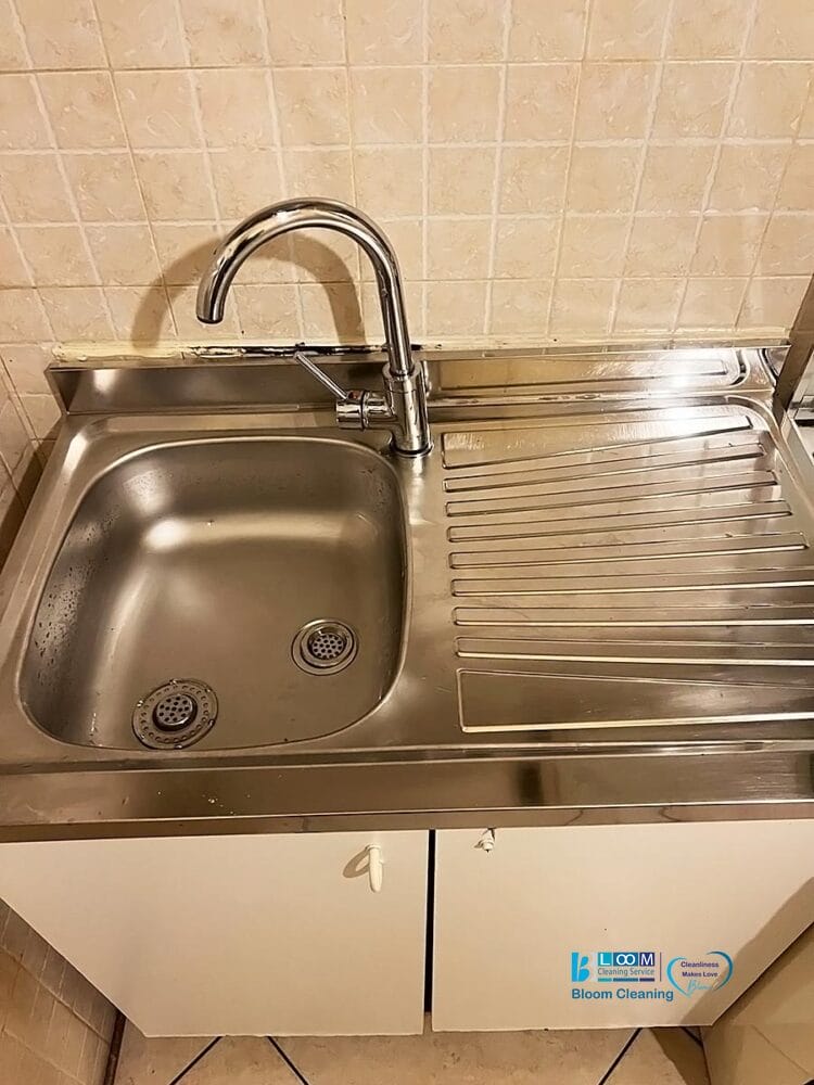 Un lavello da cucina in acciaio inox con bozza automatica, dotato di un'area per l'asciugatura e di un rubinetto singolo curvo, inserito in una parete piastrellata beige sopra mobili bianchi.