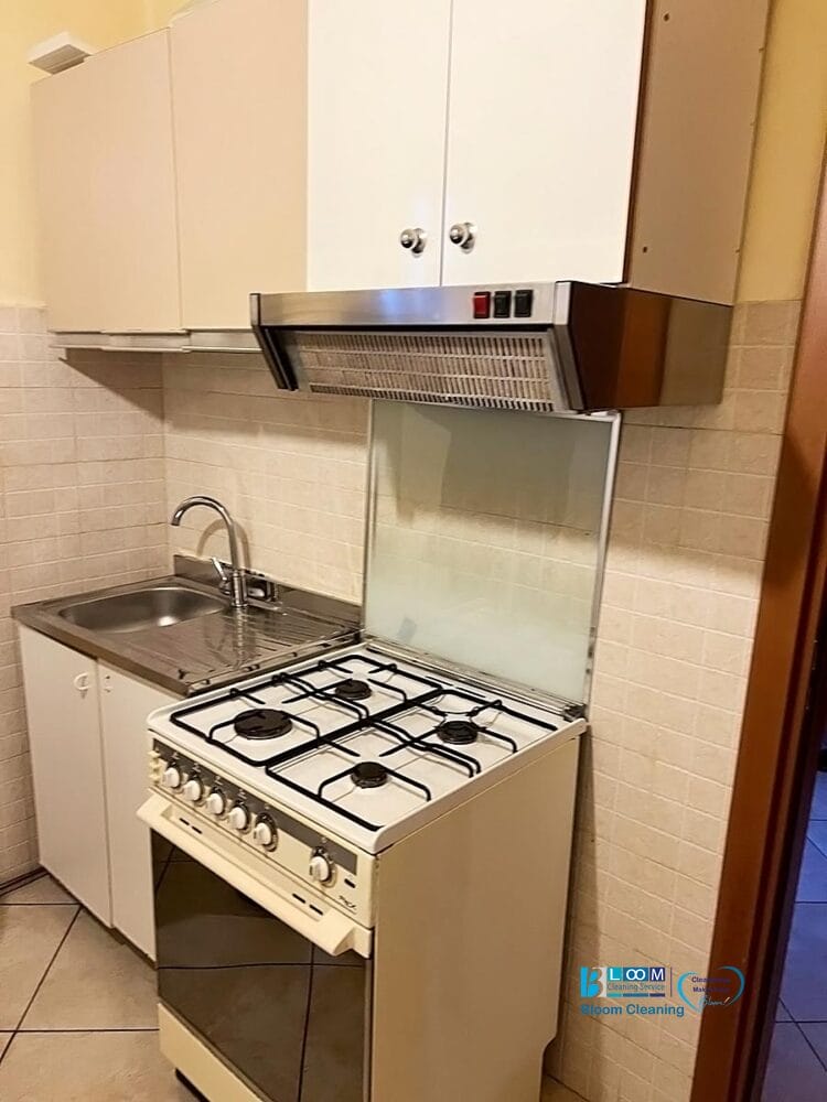 Una cucina compatta presenta un fornello bianco con quattro fuochi a gas, un forno e un backsplash trasparente accanto a un lavello in acciaio inox. Sopra i fornelli e il lavello sono montati armadietti automatici Bozza e una cappa aspirante.