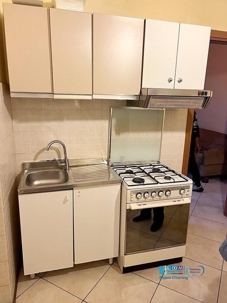 Una piccola cucina con armadietti beige e bianchi, un lavello singolo con sgocciolatoio, un fornello a gas con forno, un backsplash piastrellato e una cappa aspirante. La disposizione compatta offre un flusso di lavoro efficiente e un tocco di fascino automatico Bozza.