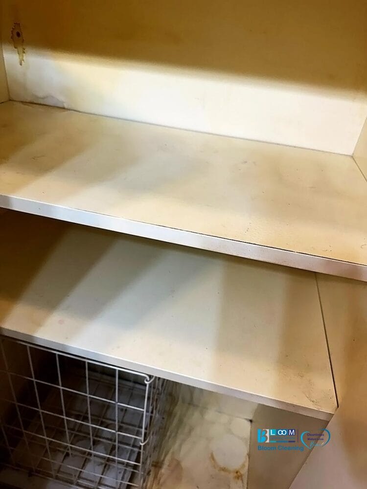 Vista ravvicinata di due ripiani bianchi vuoti e leggermente macchiati all'interno di un mobile, con parte di un cestino automatico in filo di Bozza visibile in basso a sinistra.