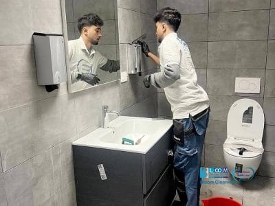 Un uomo con i guanti pulisce uno specchio sopra un lavandino in una moderna toilette con pareti piastrellate grigie. Un water con coperchio aperto e un secchio rosso sono visibili nelle vicinanze, a dimostrazione del lavoro accurato di un'impresa pulizie Garbagnate Milanese.