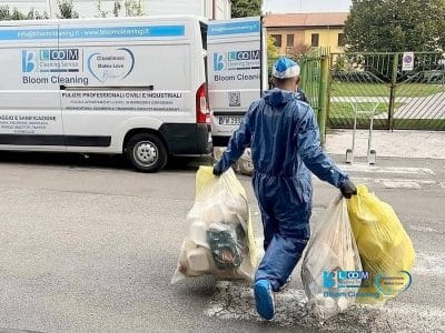 Una persona in tuta protettiva blu e berretto porta due grandi sacchi gialli di rifiuti verso un furgone del servizio di pulizia automatica Bozza parcheggiato sulla strada, con la scritta "Bloom Cleaning Service" e le informazioni di contatto.