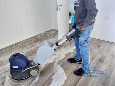 Una persona che pulisce un pavimento in legno con una macchina professionale, muovendosi sopra l'acqua saponata in una stanza per lo più vuota con pareti bianche: l'ideale per Pulizia Appartamento post ristrutturazione a Sesto San Giovanni.