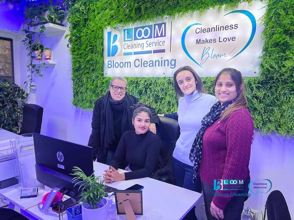 Quattro donne posano e sorridono in un ufficio moderno con una parete verde, sotto l'insegna "Bloom Cleaning Service". In primo piano c'è una scrivania con un computer e una pianta, che evidenzia le loro numerose recensioni su Google per l'eccellente servizio.