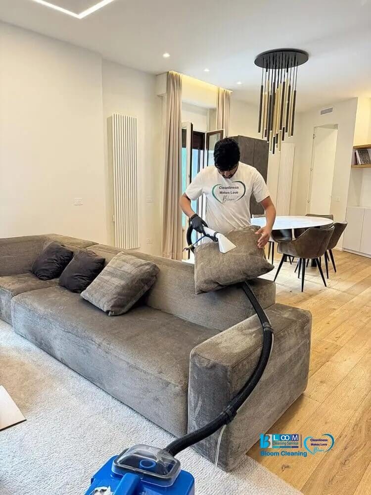 Una persona usa l'aspirapolvere per pulire i cuscini di un divano grigio in un moderno soggiorno di Milano, caratterizzato da pavimenti in legno e illuminazione elegante. La maglietta bianca mostra il logo di Pulizia appartamenti, ideale per ogni cambio inquilino.