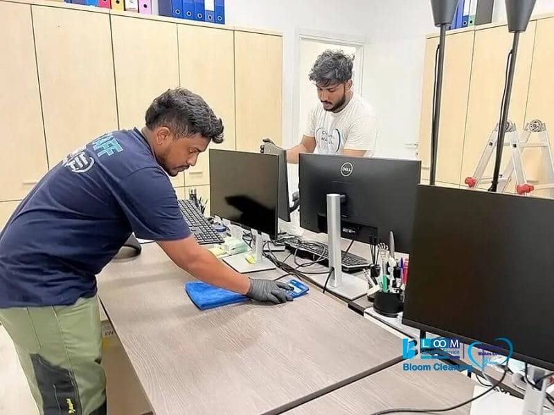Due addetti di Bloom Cleaning puliscono una scrivania d'ufficio con monitor di computer nella Provincia di Milano, uno pulisce la superficie con un panno in microfibra blu mentre l'altro riordina le attrezzature vicine nell'ambito dei nostri Servizi Multiservice.
