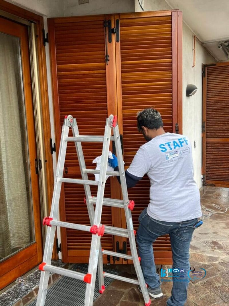 Una persona con la maglietta "STAFF" è in piedi su una scala a pioli per pulire o lavorare sulle persiane in legno attaccate a un edificio, che mostra la competenza multiservizi di Milano e Provincia, con altre porte in legno e un pavimento piastrellato a vista.